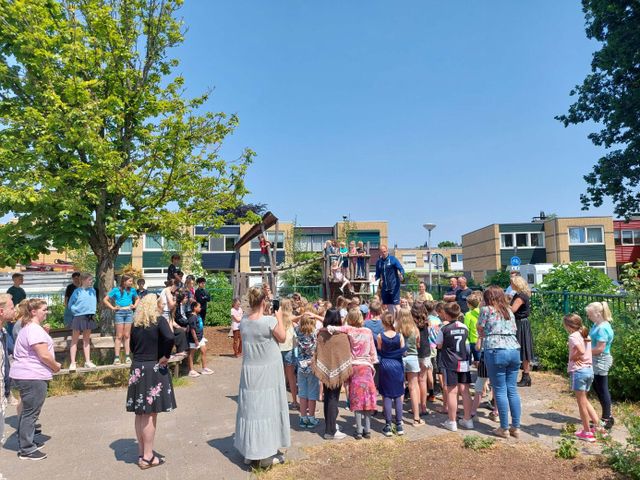 Een meneer op een plein verteld wat aan kinderen en ouders met op de achtergrond een speeltoestel en huizen.