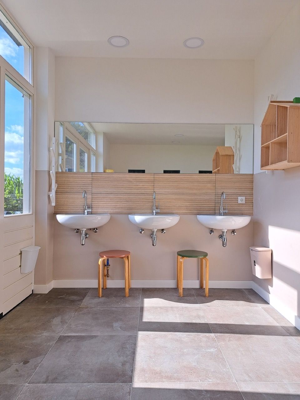 Drie witte wasbakken met kranen aan de muur in een lichte sanitaire ruimte, met twee houten krukjes ervoor en een grote spiegel erboven.