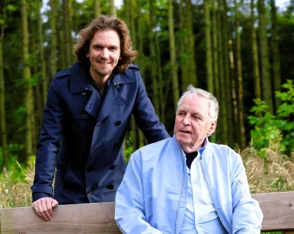Twee mannen, De oudere man zit op een houten bankje en kijkt opzij, terwijl een jongere man naast hem staat en zijn hand op de rugleuning legt. Beiden dragen jassen. De foto straalt rust en onderlinge verbondenheid uit.