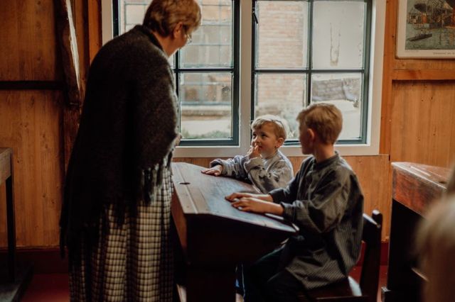 Een vrouw in klederdracht vertelt in een schooltje iets aan twee kinderen