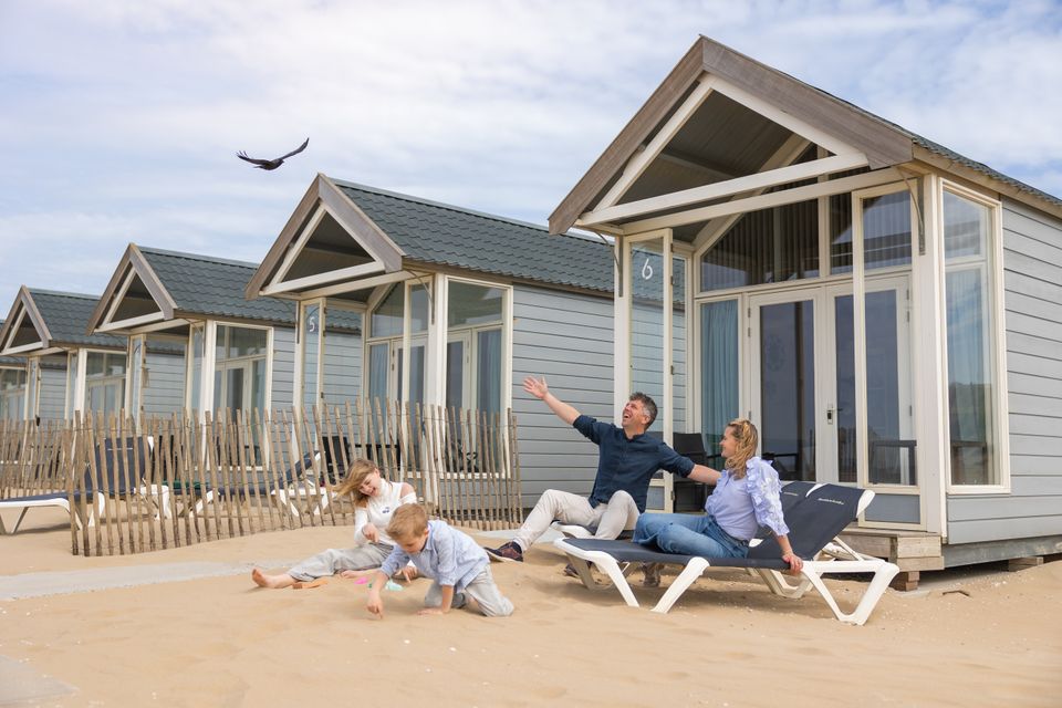Familie van vader, moeder en twee kinderen zit blij voor een strandhuisje in Katwijk, een meeuw vliegt over.