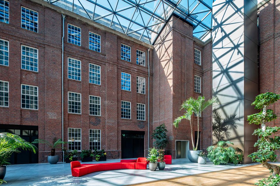 Foto van een met glas overdekte open ruimte (atrium). Zichtbaar zijn de ramen van de oude leerfabriek in de gevel, rode banken en groene planten in het midden.