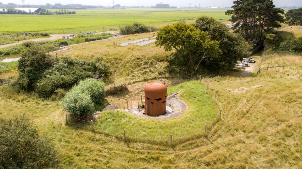 De cortenstalen vogelobservatiekoepel op Fort K'ijk. Er omheen zie je bomen, gras en mensen op een terras.