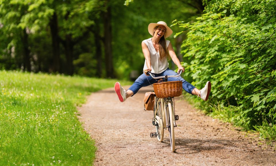 Vrouw fietst vrolijk over een groen bospad met haar benen speels omhoog, op een fiets met rieten mandje, omringd door bomen en zomerse natuur.
