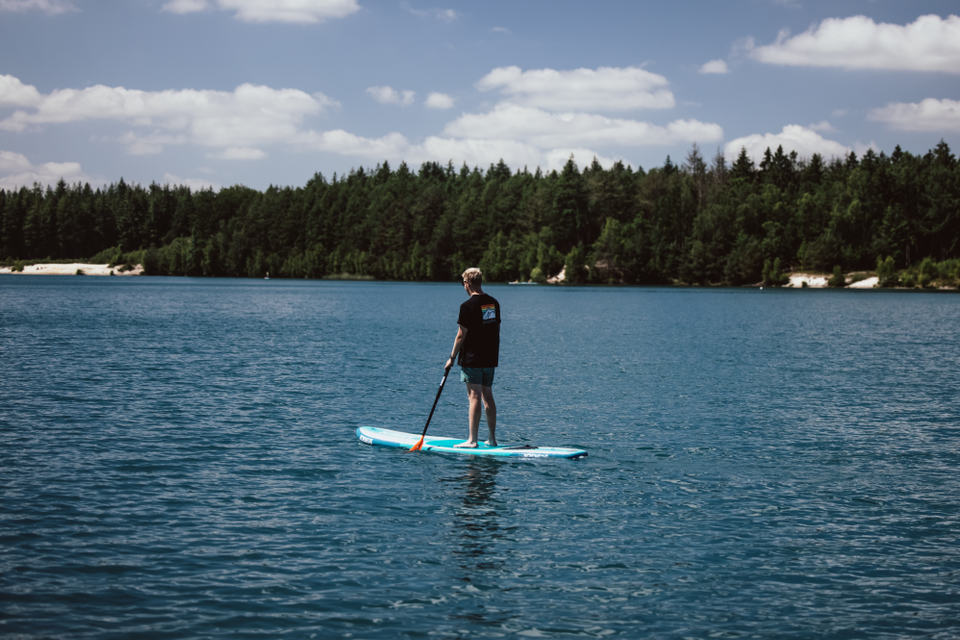 Ein Mann steht auf einem SUP-Board und paddelt über blaues Wasser vor einer Kulisse aus Nadelbäumen.