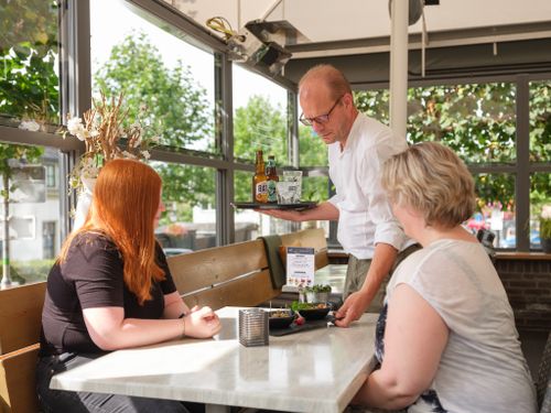 Twee vrouwen zitten aan een tafel in een restaurant, een man serveert hapjes en drankjes.