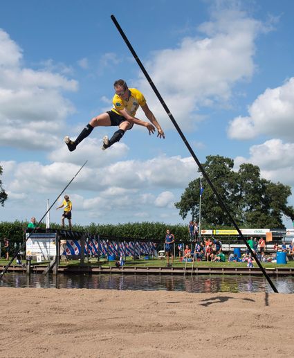 Man springt met een polsstok over het water tijdens het NK Polsstokverspringen, met toeschouwers op de achtergrond in Linschoten.