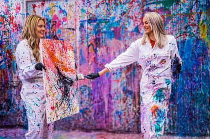 Twee vrouwen in witte overalls maken een kleurrijk schilderij door verf te spatten tijdens de creatieve activiteit Gooien met Verf bij Fabriek Woerden.