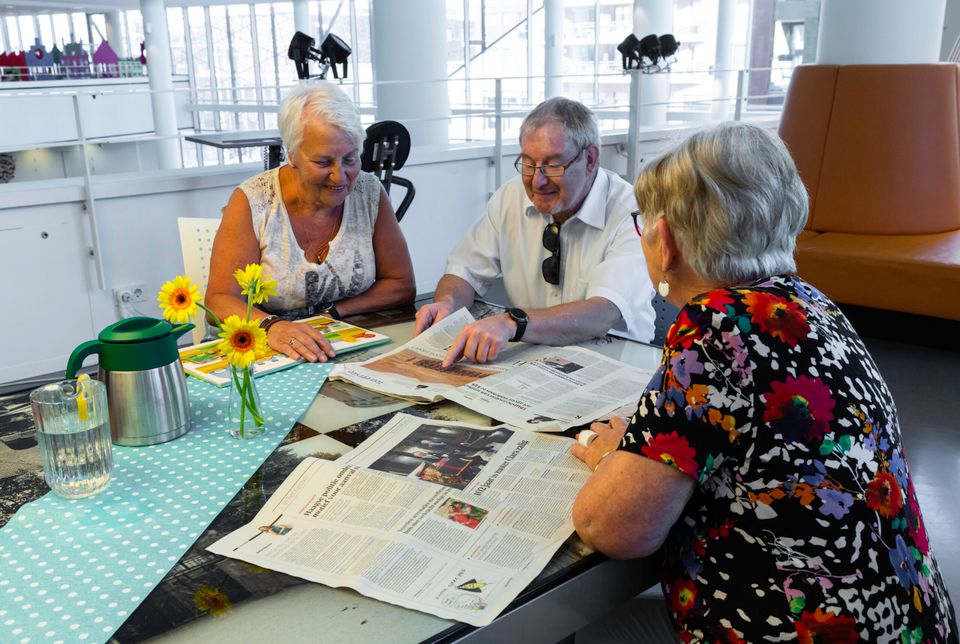 Op de foto zitten een aantal mensen aan een tafel, ze hebben kranten en boek voor zich en wijzen naar een foto in de krant. Op de tafel staan gele bloemen in een vaas en een kan water en een kan koffie.