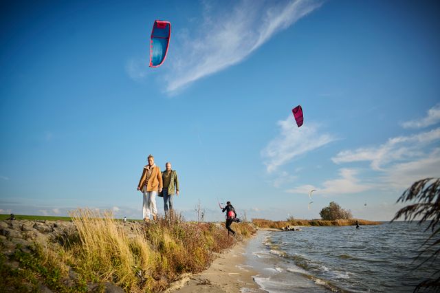 Twee mensen wandelen achter elkaar langs het IJsselmeer bij het Mirnser Klif met kites in de lucht.