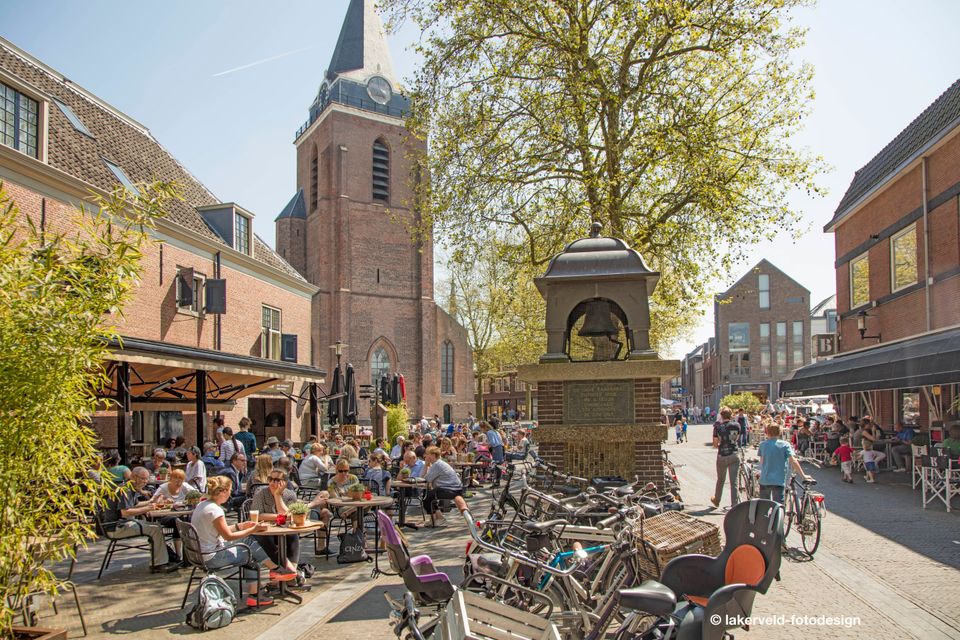 Zicht op de Kaasbel, Petrustoren en het Kerkplein te Woerden met volle terrasjes, fietsen op de voorgrond en wandelende mensen over het plein.