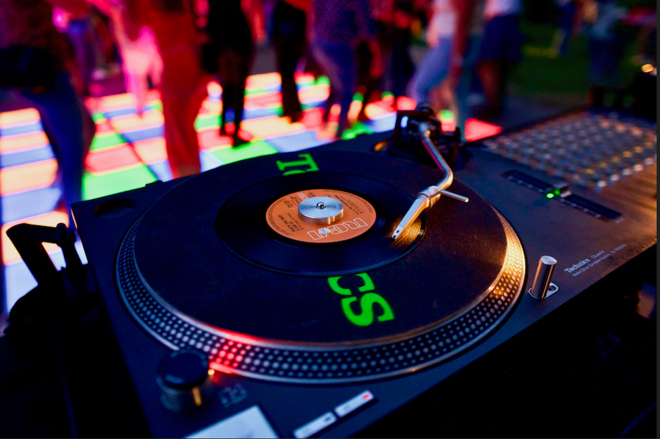 Foto van een draaitafel met op de achtergrond een kleurrijke verlichte dansvloer.
