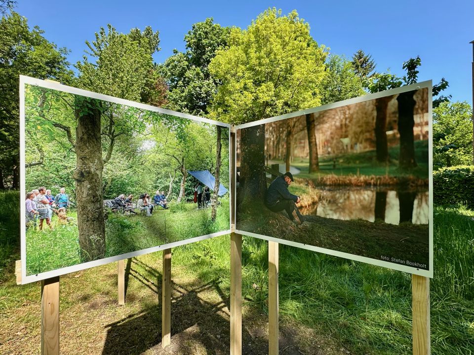 Twee grote buitenfoto’s op houten panelen op Landgoed Bredius in Woerden, waarop links een groep mensen in een bosrijke omgeving te zien is en rechts een man zittend bij de waterkant, onder heldere blauwe lucht.