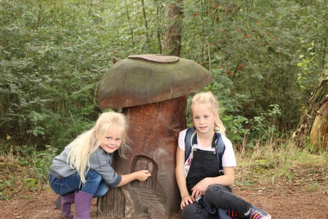twee blonde meisjes bij een houten beeld van een paddenstoel in het bos