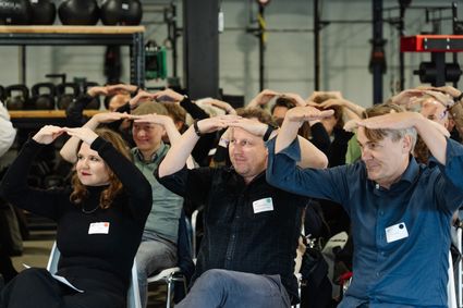 Mensen zitten in zaal en houden beide handen plat boven het hoofd.