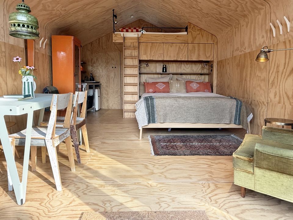 Interieur van een wikkelhuisje bij Proeftuin van Linschoten met houten wanden, tweepersoonsbed met opbergruimte, eettafel met stoelen, kitchenette en een trap naar een slaapvide.