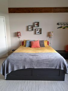 Slaapkamer met tweepersoonsbed, gele sprei en grijze deken, afgewerkt met kleurrijke kussens, wandlampen en kleine wandplanken boven het hoofdbord.