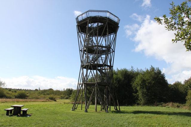 Een uitkijktoren midden in een open veld. Voorin staat een picknickbank