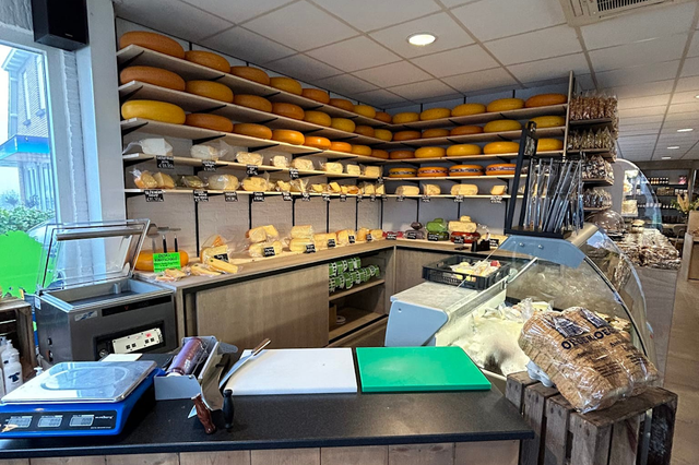 Een foto van een toonbank met op de achtergrond planken waar hele kazen op liggen.