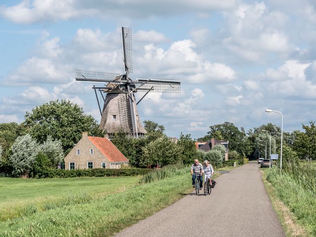 twee mensen die langs een molen fietsen, naast de molen staat een klein huisje.