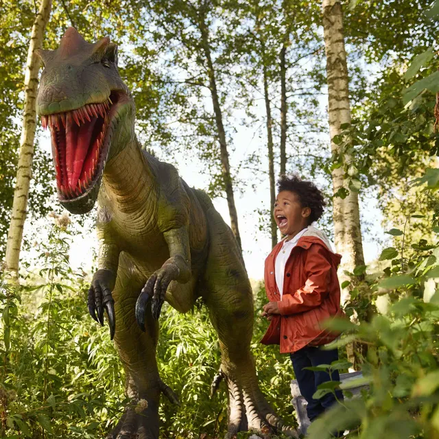 Een kindje die met een grote lach naast een levensgrote dino staat.