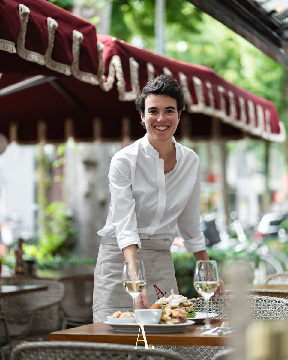 Serveerster in witte kleding op terras van restaurant AMBIANS. in Oisterwijk onder bordeauxrode parasol