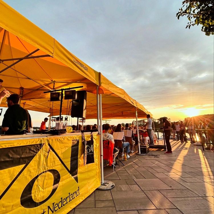 Gele open tent waarin mensen zitten die instrumenten bespelen. Op de achtergrond staan mensen en is de avondzon te zien.