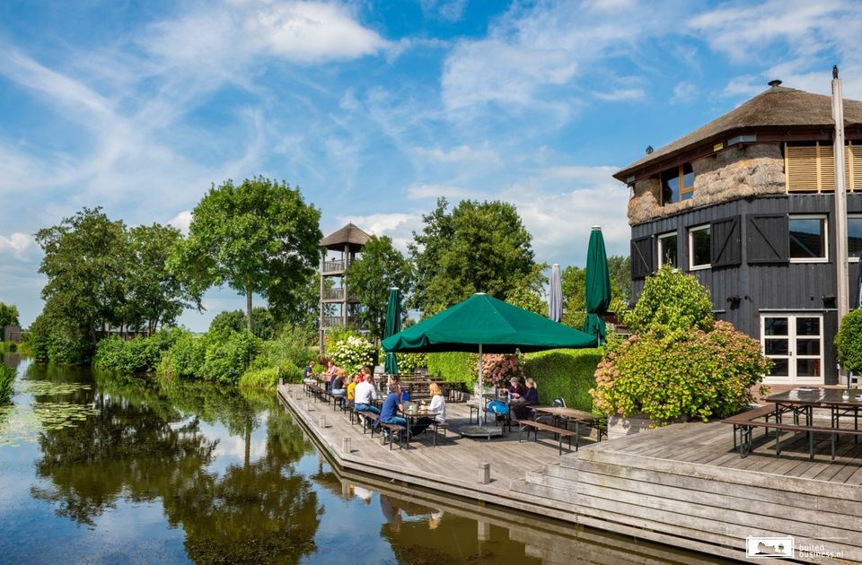 Restaurant met terras Boerderij De Boerinn in Kamerik, Groene Hart. Landelijke horecalocatie aan het water met ruim houten terras, gasten die buiten zitten, rietgedekt gebouw, groen polderlandschap en uitkijktoren op een zonnige dag.