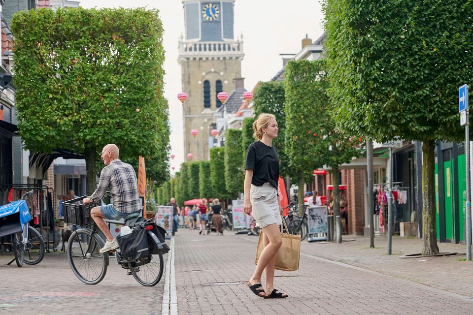 Een fietsende man en winkelende vrouw met boodschappentas lopend op de Midstraat in Joure. Met op de achtergrond de kerktoren en in de lucht Chinese lampionnen.