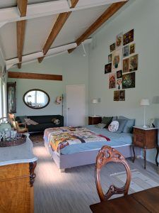 Lichte slaapkamer met twee eenpersoonsbedden, houten plafondbalken, nachtkastjes en wanddecoratie, verlicht door grote ramen en een ronde spiegel aan de muur.