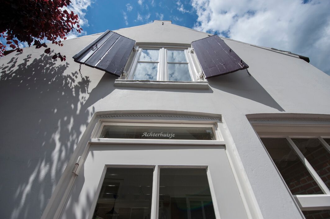Witte gevelfoto van onderaf genomen met de voordeur en een raampje erboven met rode luiken. Boven de voordeur in het glas staat "Achterhuisje" gegraveerd.