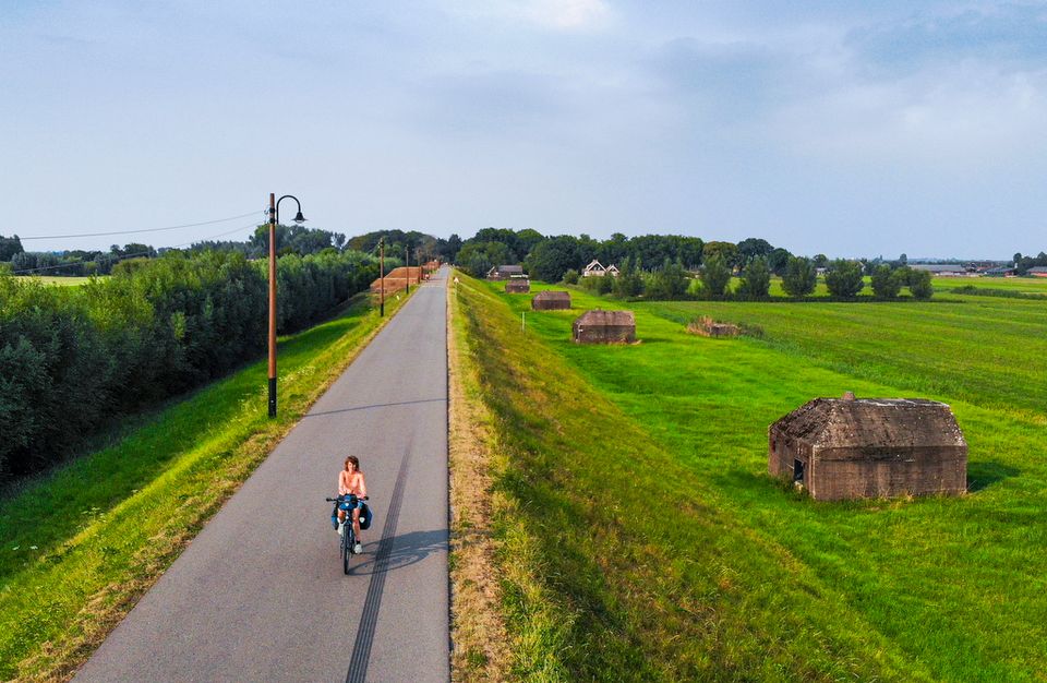 Een fietster fietst over een asfaltpad op een dijk. Langs de dijk liggen verschillende betonnen groepsschuilplaatsen.