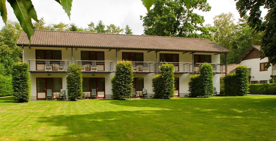 Witte gevel met de diverse kamers met balkon op een groot groen grasveld en op de achtergrond wat bomen.