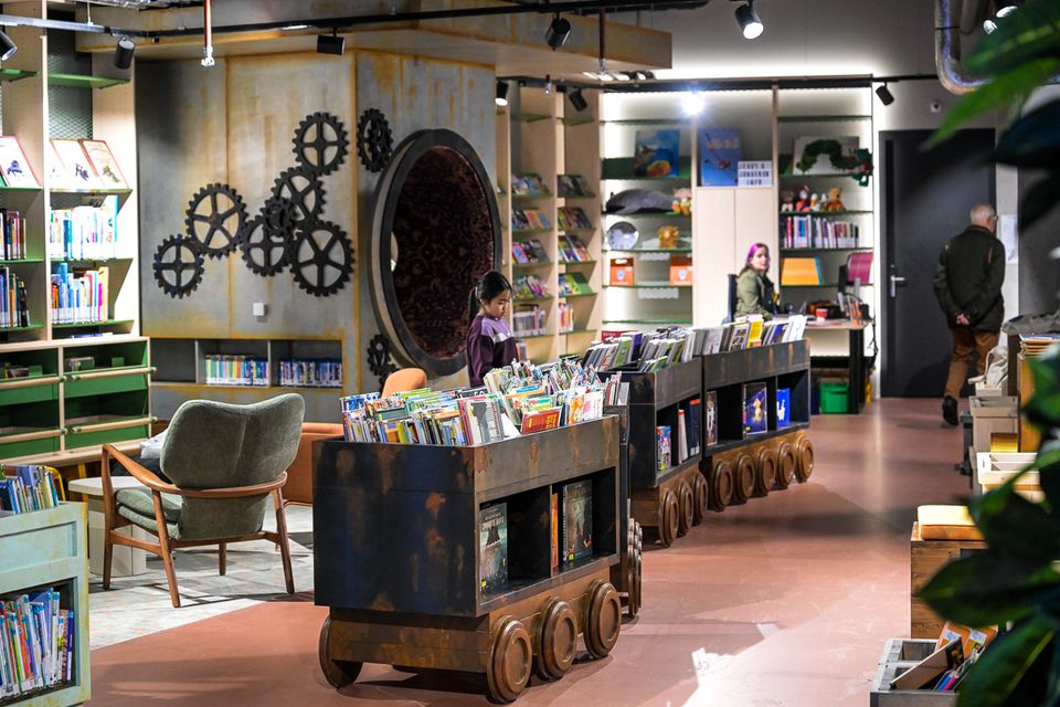 Interieur van een moderne bibliotheek met verrijdbare boekenkarren gevuld met kinderboeken. Bezoekers lopen en lezen tussen de kasten. De ruimte heeft industriële elementen, zoals metalen details en decoratieve tandwielen aan de wand.