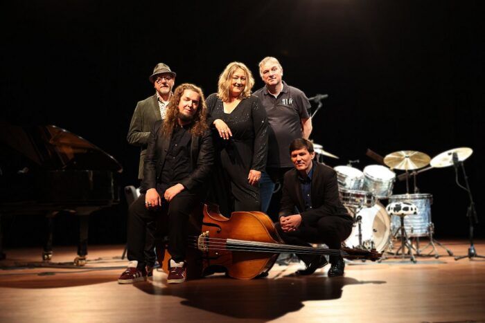 Vijf muzikanten poseren op een podium met instrumenten, waaronder een contrabas, piano en drumstel. Ze dragen donkere kleding en kijken richting de camera. De setting en belichting duiden op een bandfoto voorafgaand aan een optreden.