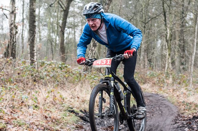 Man in blauwe jas met bril en helm op op een mountainbike door het bos. Hij gaat van een heuvel af en lijkt snel te gaan.