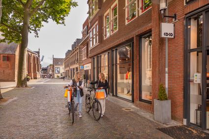 Winkelgebied in de binnenstad van Woerden, Groene Hart, waar twee vrouwen met fietsen en volle winkeltassen langs winkels in het historische centrum lopen.