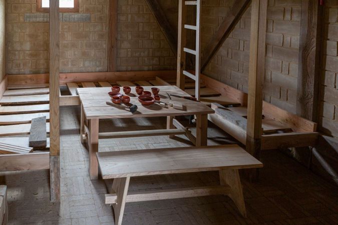 Houten banken en tafels in de soldatenbarak, met op tafel aardewerk drinkgerei.