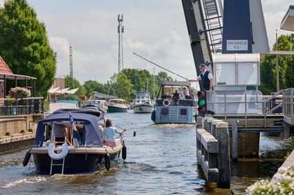 Er vaart een jacht met een man en een vrouw erop onder de open brug van Delfstrahuizen door en er wordt drie euro aan bruggeld betaald aan de brugwachter doormiddel van een hengel met een klomp. Op de achtergrond zijn varende en vastliggende boten te zien.