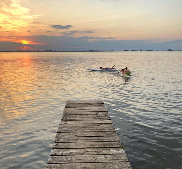 Kinderen op SUPboards bij een steiger met een laaghangende zon.