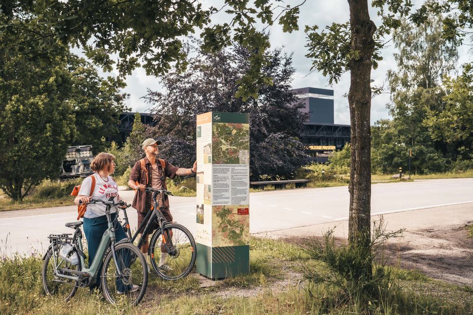 Twee personen met fiets in hand kijken naar Toeristisch Oversap Punt (informatiebord). Ze staan bij het Nationaal Militair Museum in Soest in regio De Utrechtse Heuvelrug.