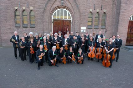 Culturele muziekuitvoering Het Groene Hart Orkest in Woerden, Groene Hart, groepsfoto van muzikanten met violen, cello’s en blaasinstrumenten voor bakstenen kerkgebouw tijdens concert of repetitie.