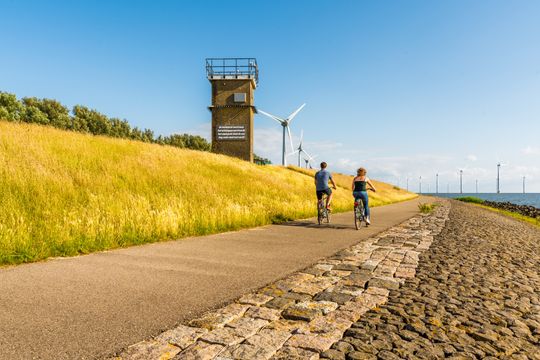 Man en vrouw fietsen over de dijk langs Rotterdamse Hoek in de Noordoostpolder met aan hun Linkerzijde de dijk en aan hun rechterzijde water en windmolens.