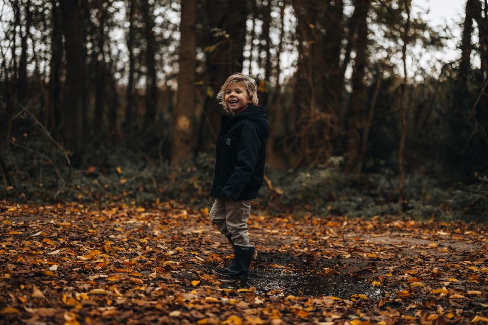 Een jongetje staat op een herfstdag in het bos met zijn laarzen in de modder.