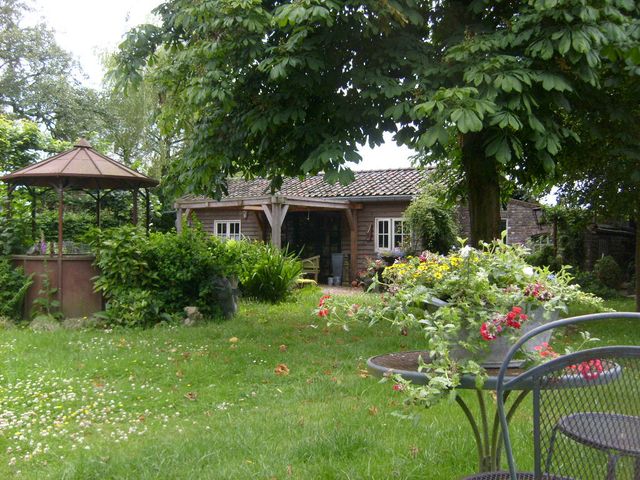 Zicht op een houten chalet met afdak op een groen grasveld met aan de linkerkant een 6-hoekige volière en op de voorgrond rechts een smeedijzeren zitje met kleurrijke bloemen.