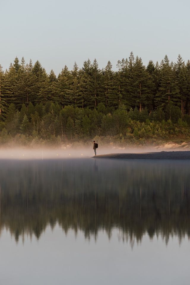 Een man staat in de verte bij het water waar een laag mist boven hangt.