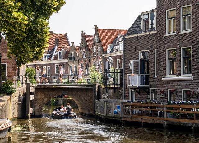 Een sloep vaart door een smalle gracht onder een lage brug, terwijl wandelaars de brug oversteken. Langs het water staan historische huizen met trapgevels en terrassen, omgeven door groen, in de binnenstad van Oudewater.