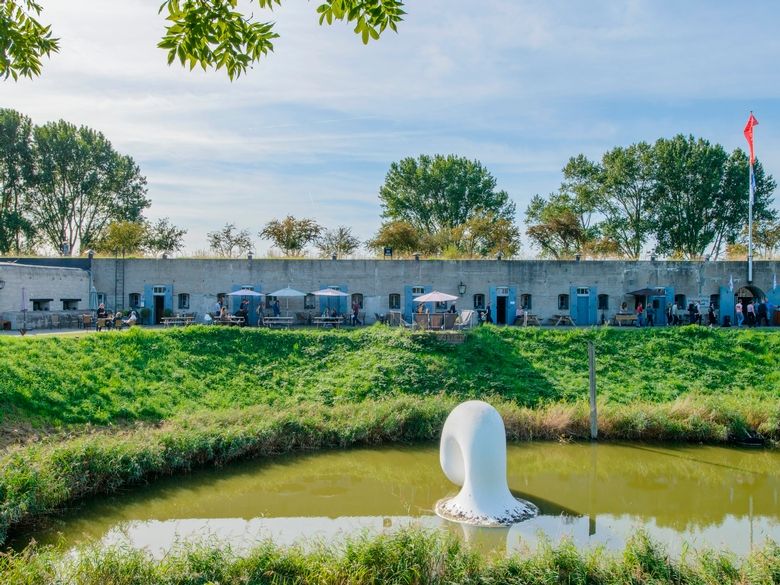 Een betonnen fort met daarvoor een fortgracht. In de gracht is een wit, kunststof kunstwerk geplaatst.