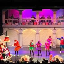 Foto van een podium met in de linker beneden hoek Sinterklaas in zijn stoel en op het podium pieten die hem toezingen.