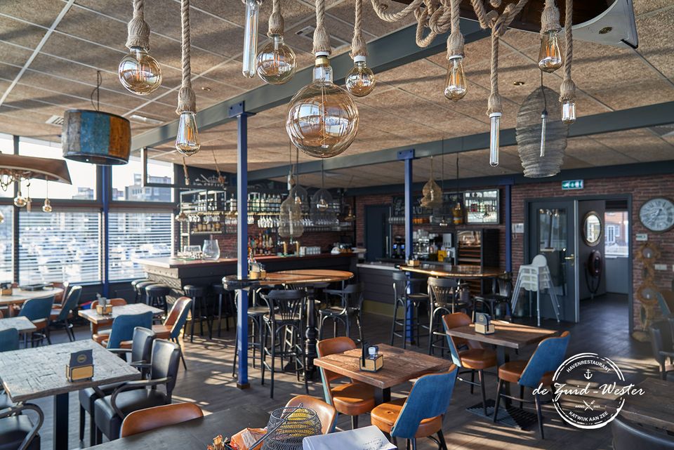 Sfeerfoto van Havenrestaurant De Zuid-Wester, met tafels en stoelen, een kleine bar en scheepstouwen met moderne lampen aan het plafond.